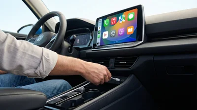 Close-up view from the passenger seat of a person connecting a smartphone via USB-C cable to the dashboard console of a modern rental car, with a brightly lit infotainment screen.