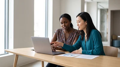 Two women compare on-airport and off-airport car rental costs for New York in a bright office.