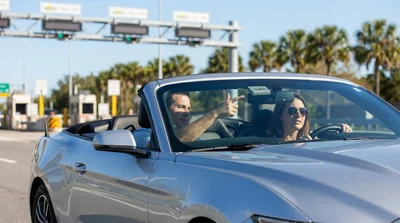 Silver car rental driving under a highway toll plaza on a sunny day in Orlando