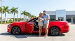 Two friends exchanging keys next to a modern car rental parked on a sunny palm-lined street in Florida