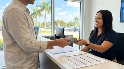Traveler holding a credit card and passport next to a car rental vehicle parked under palm trees in Florida