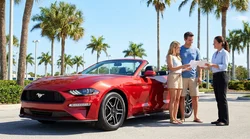 Two people driving a white car rental along a sunny coastal road with palm trees in Florida