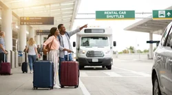 Passengers with luggage boarding a white car rental shuttle bus outside an airport terminal in Pennsylvania