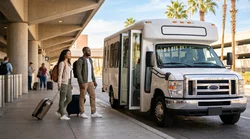 Passengers loading luggage into a white car rental shuttle bus parked outside a Las Vegas airport terminal