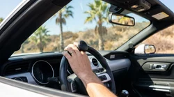Driver's hands using a spinner knob on a car rental steering wheel with a sunny California road visible outside