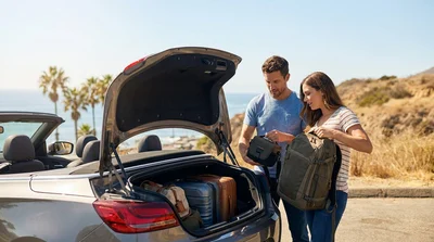 Traveler loading luggage into a car rental trunk parked along a sunny California coastline