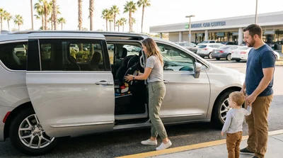 Parent buckling a toddler into a child seat inside a car rental parked on a sunny Los Angeles street