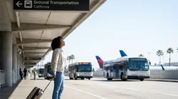 Passengers with luggage boarding a car rental shuttle bus outside a busy Los Angeles airport terminal