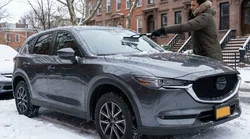 Person using an ice scraper to clear snow from a car rental parked on a winter street in New York