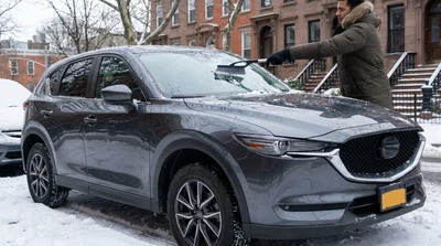Person using an ice scraper to clear snow from a car rental parked on a winter street in New York