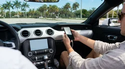 Driver plugging a smartphone into a dashboard screen inside a car rental on a sunny Miami street