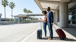 Travelers with luggage waiting for a car rental shuttle outside a Los Angeles airport terminal