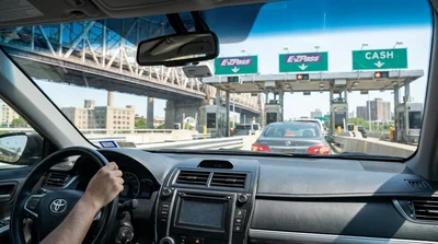 A silver car rental approaching a New York bridge toll booth under a clear blue sky