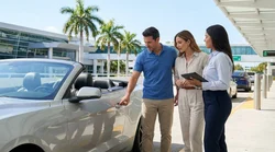 Person inspecting a scratch on the bumper of a white car rental parked near palm trees in Miami