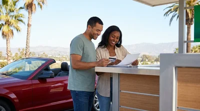 A smiling couple driving a car rental along a sunny coastal highway in California