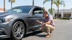 Driver checking the bumper of a parked car rental for scratches on a sunny street in California