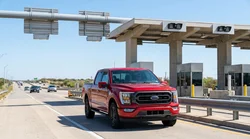 A silver car rental driving on a multi-lane Texas highway under a toll road gantry