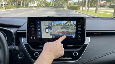 Dashboard screen displaying a rear-view camera inside a car rental parked near palm trees in Florida