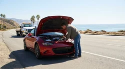 Driver looking under the hood of a broken-down car rental on the shoulder of a sunny California highway