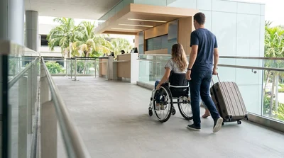 Traveler with rolling luggage on a flat step-free path approaching a car rental facility in Miami