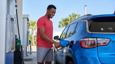 A man happily fills the fuel tank of a modern blue Jeep Compass at a gas station under bright Orlando daylight.