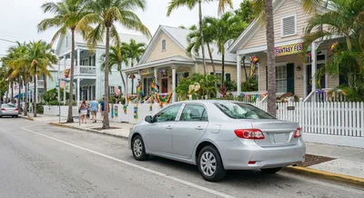 A silver rental car parked on a quaint, palm-lined residential street in Key West featuring colorful tropical architecture.