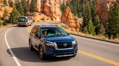 Couple driving a blue Nissan SUV rental in a national park with a guided tour bus in the background.