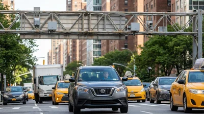 A grey rental car driving under toll sensors on a busy New York City street during the day.