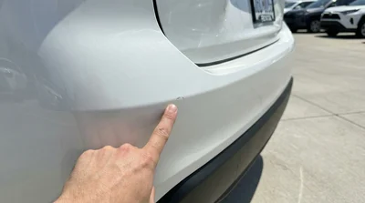 Close-up shot of a person's hand pointing to a small, minor scuff mark on the rear white bumper of a Toyota Rav4 rental car during an exterior inspection.