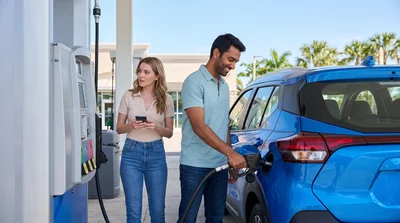 A diverse couple refuels a blue Nissan Kicks rental car at a Florida gas station in bright daylight.