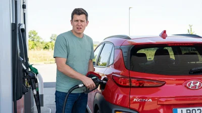 A person looks confused while holding a gas pump nozzle next to a red Hyundai Kona car rental.